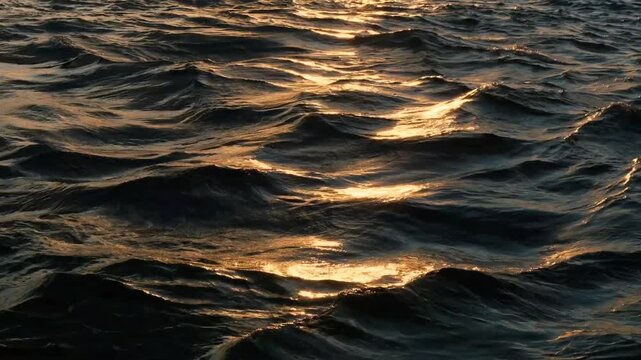 Golden hour sun glare on dark sea surface water ripples. Close up of warm orange sunset light shimmering over mysterious deep blue ocean waves at evening.