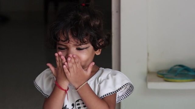 Close-up portrait of a cute little Indian toddler girl standing indoors, playfully rubbing her hands together and smiling warmly at the camera with an innocent expression.
