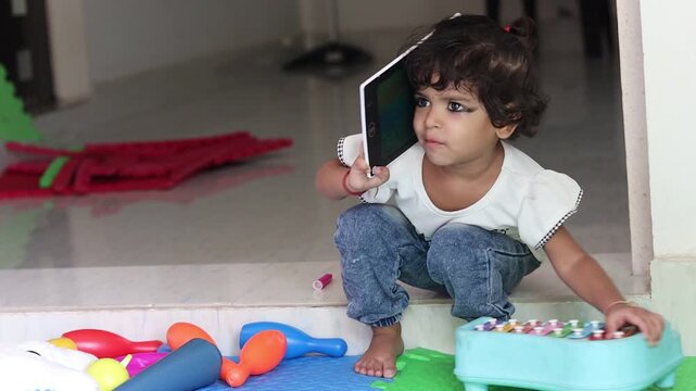 A cute little Indian toddler girl sitting indoors near scattered toys, playfully pretending to talk on a toy tablet like a mobile phone, showing early childhood imagination and pretend play.
