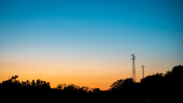 Dramatic sunset over a dark silhouette of trees and a high voltage power pylon. Orange and blue twilight sky with electrical lines. Concept for energy, industry, power distribution, and nature.