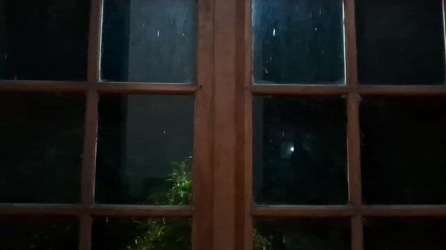Raindrops fall at night when hit by a spotlight, visible from behind a wooden-framed glass window