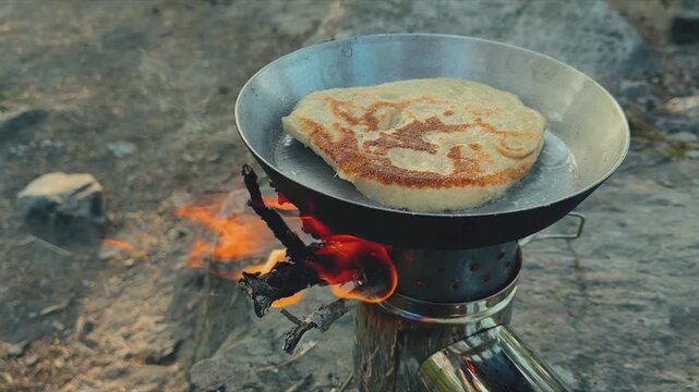 Flatbread frying in steel pan on wood-burning camping stove over open flame, outdoor survival cooking