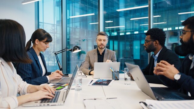 Business team working at table with laptops and charts, leader pointing while discussing strategy. Focused professionals in formal attire engaged in project planning in modern office.