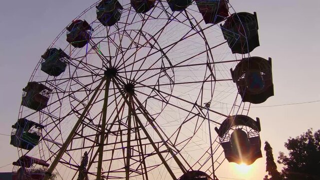 Silhouetted Ferris wheel spins against twilight sky. Sun sets behind amusement park ride, casting warm glow. Passengers ride, suspended in slow, rhythmic motion. Structure stands tall