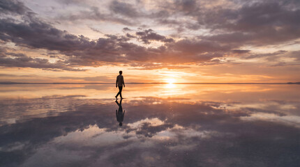 Obraz premium Flooded salt flat at golden hour with perfectly flat water reflecting dramatic cloudy sky as one person walks across the surface appearing to walk through clouds