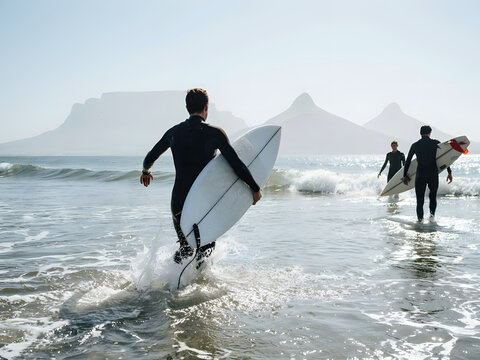 Surfers running into ocean waves with surfboards, beach scene with mountains