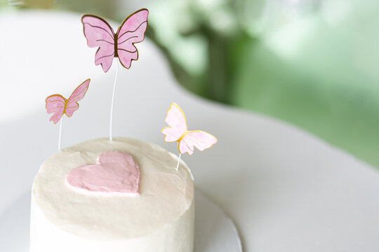 Small white cake with pink heart and butterfly decorations on a table, pastel dessert styling, romantic celebration, sweet party mood.