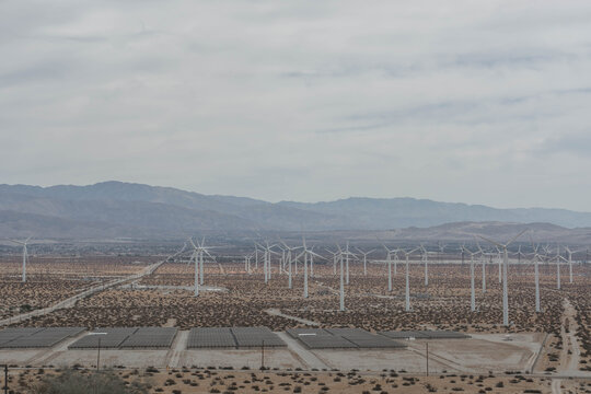 High angle overview of a vast wind farm and solar panel arrays stretching across a flat desert valley toward distant hazy hills.