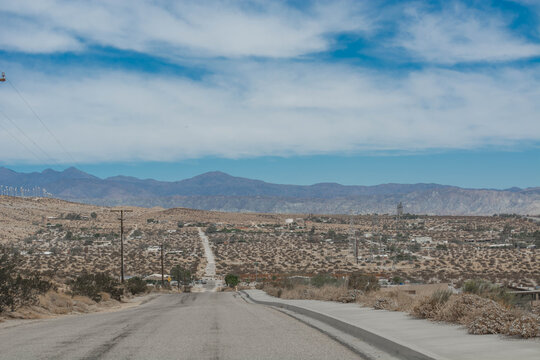 A long empty asphalt road stretching down through a dry desert landscape toward a distant blue mountain range under a cloudy sky.