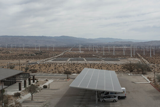 Aerial view of a solar panel canopy over a parking lot in a desert facility surrounded by massive wind turbine fields.