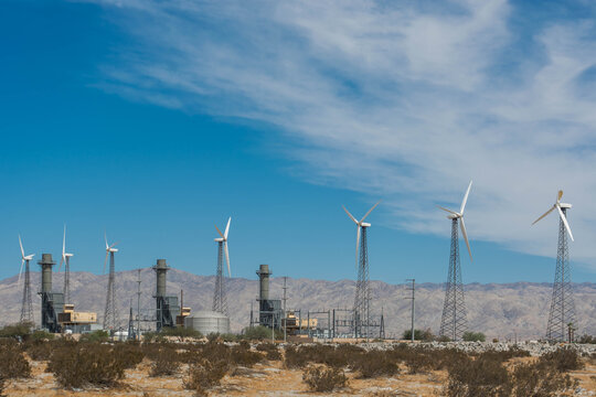 Traditional lattice style wind turbine towers standing in a row in a desert facility with power plant structures and distant mountains.