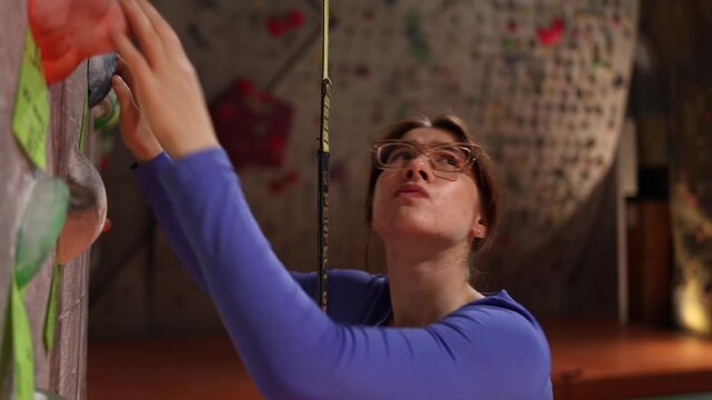 Beginner woman on indoor climbing wall hesitates before next move looking up with tension and concentration showing fear uncertainty and decision momentBeginner woman on indoor climbing wall hesitates
