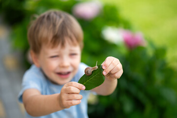 Happy little boy holding green leaf with garden snail during outdoor nature exploration. Child curiosity, discovery and learning in lush blooming garden. Outdoor adventure carefree summer childhood. © Тетяна Немировська
