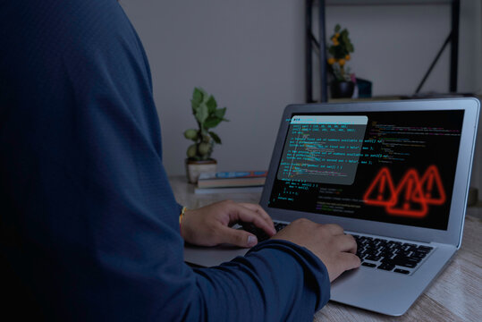 Over the shoulder view of a programmer typing on laptop with source code and glowing red warning icons on screen. Dark indoor lighting, software bug debugging and system error concept.