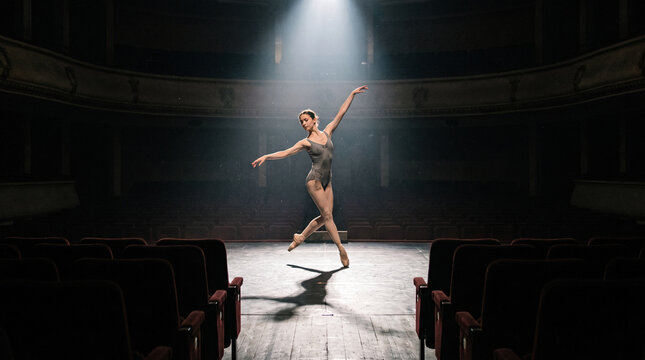 Young ballerina dancing alone on large 
empty stage illuminated by single spotlight 
with vast dark theater seating completely 
empty beyond the stage edge
