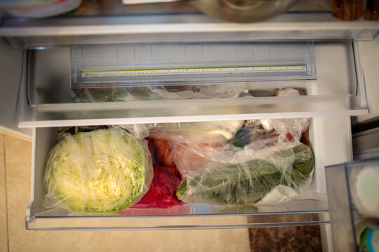 Open refrigerator vegetable drawer with fresh cabbage in plastic bag, greens and vegetables stored inside, cherries and strawberries visible above