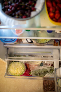 Open refrigerator vegetable drawer with fresh cabbage in plastic bag, greens and vegetables stored inside, cherries and strawberries visible above