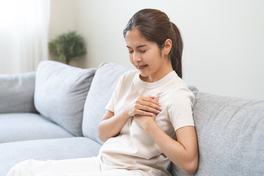 Heart attack, health care concept, suffering acute cardiac asian young woman hand touching on her chest pain, female breathing problem, asthma attack, pressure while sitting on sofa, couch at home.