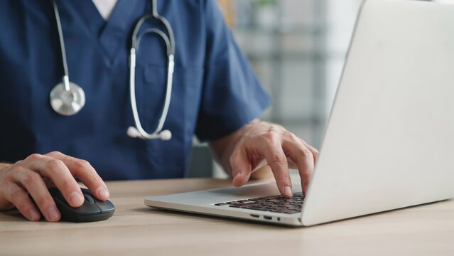 A close-up photo of a medical professional in blue scrubs with a stethoscope around their neck, typing on a silver laptop and using a black computer mouse on a wooden desk.