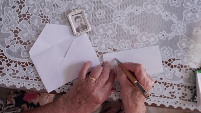 Mature woman's wrinkled hands writing a romantic letter with a pen on a lace tablecloth, a small silver framed picture of a young man nearby, expressing nostalgia, memory, and enduring love