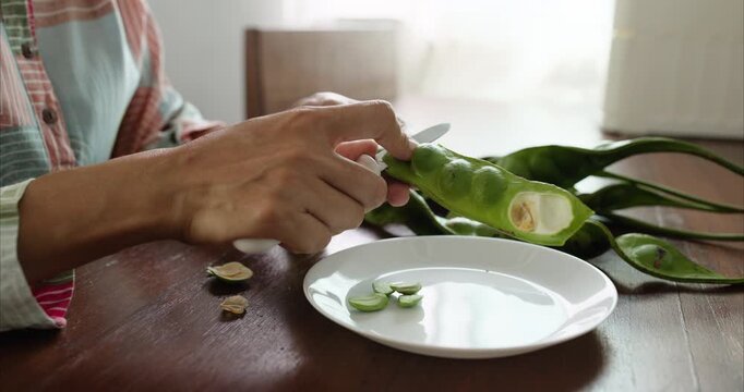 Close up. Asian Woman Hand Peeling Parkia Speciosa , Stink Bean, Bitter Bean, with Knife into White Plate on Table.