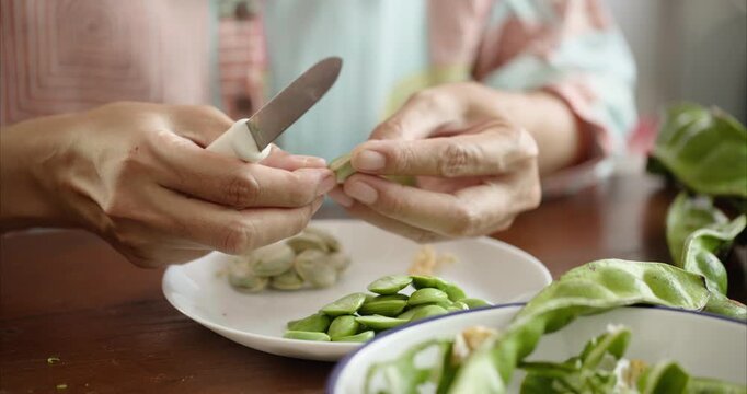 Close up. Asian Woman Hand Peeling Parkia Speciosa , Stink Bean, Bitter Bean, with Knife into White Plate on Table.