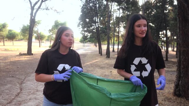 Dedicated female activists wearing recycling symbol t shirts and gloves walk through a park, holding a green trash bag open to collect garbage and combat environmental pollution together