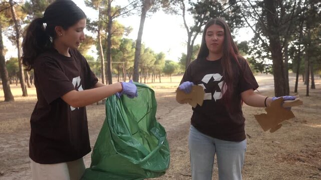 Two dedicated young women wearing recycling symbol t shirts and gloves actively collect and sort cardboard waste into a green garbage bag during a park cleanup