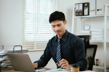 Fototapeta premium Young businessman working on a laptop in a modern office, reviewing documents and business reports during a productive workday.