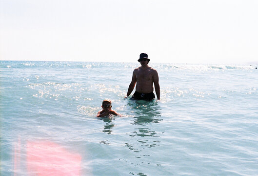Child Swims Near Adult in Shallow Water at Beach During Sunny Day
