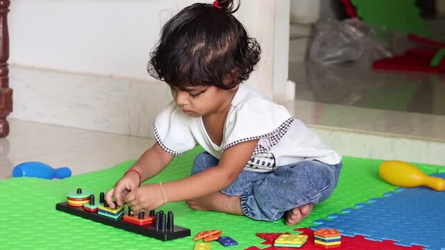 A cute little Indian toddler girl sitting on a colorful playmat indoors, deeply engaged in activity-based learning by sorting colorful wooden shapes on a pegboard puzzle.

