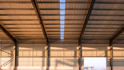 Modern empty warehouse interior with metal sheet roof and steel structure under golden evening sunlight in construction site © Prapat