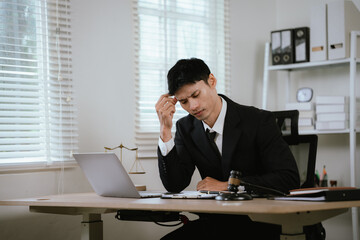 Obraz premium Stressed lawyer working on a laptop in a modern office, reviewing legal documents with a judge's gavel, symbolizing legal pressure.
