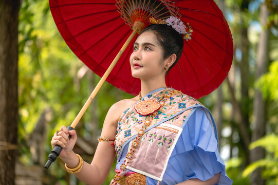 Thai woman in elegant traditional national costume and exquisite jewelry accessories , holding red paper parasol enjoying inside ancient temple in Chiang Mai
