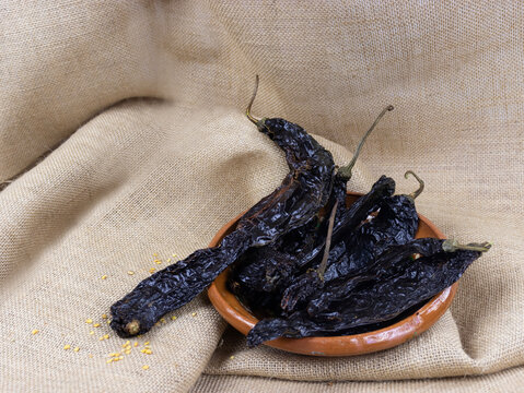 Dried pasilla chili peppers in a rustic clay bowl on a burlap background.