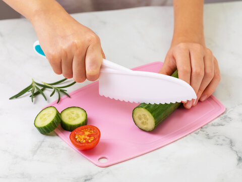 Child's hands cutting cucumber with a safe plastic knife on pink cutting board with tomato and rosemary close-up
