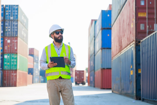 Portriat handsome male hispanic latin Industrial and factory Specialist. beard man worker wearing white protective hard hat helmet working on tablet computer at container yard.