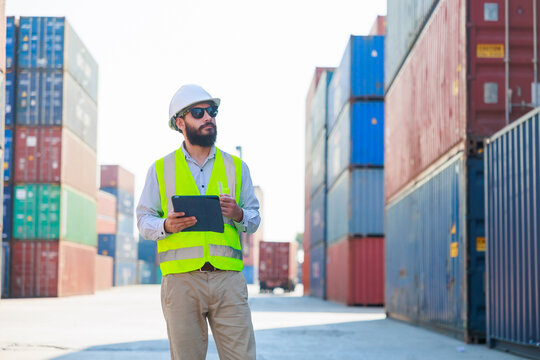 Portriat handsome male hispanic latin Industrial and factory Specialist. beard man worker wearing white protective hard hat helmet working on tablet computer at container yard.