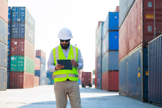 Portriat handsome male hispanic latin Industrial and factory Specialist. beard man worker wearing white protective hard hat helmet working on tablet computer at container yard.