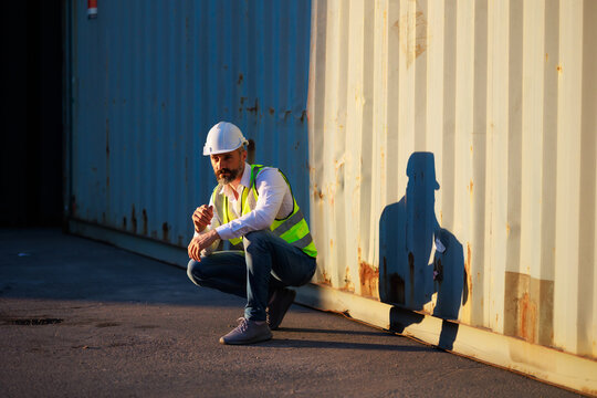 Handsome caucasian male dock foreman worker in white hard hat helmet and high-visibility vest working in Shipping Cargo Container Terminal Depot