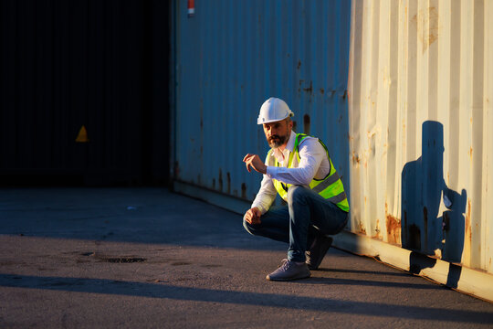 Handsome caucasian male dock foreman worker in white hard hat helmet and high-visibility vest working in Shipping Cargo Container Terminal Depot