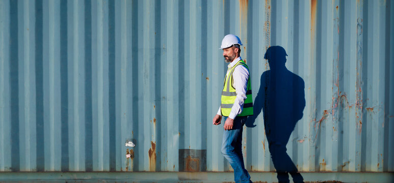 Male walk. Handsome caucasian male dock foreman worker in white hard hat helmet and high-visibility vest walking in Shipping Cargo Container Terminal Depot