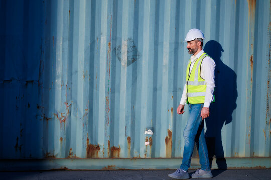 Male walk. Handsome caucasian male dock foreman worker in white hard hat helmet and high-visibility vest walking in Shipping Cargo Container Terminal Depot