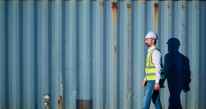 Male walk. Handsome caucasian male dock foreman worker in white hard hat helmet and high-visibility vest walking in Shipping Cargo Container Terminal Depot