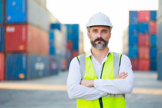 Man manager cross arm. Handsome caucasian male dock foreman worker in white hard hat helmet and high-visibility vest walking in Shipping Cargo Container Terminal Depot