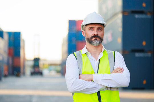 Man manager cross arm. Handsome caucasian male dock foreman worker in white hard hat helmet and high-visibility vest walking in Shipping Cargo Container Terminal Depot