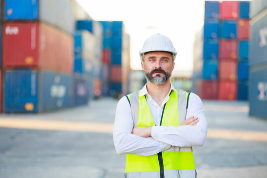 Man manager cross arm. Handsome caucasian male dock foreman worker in white hard hat helmet and high-visibility vest walking in Shipping Cargo Container Terminal Depot