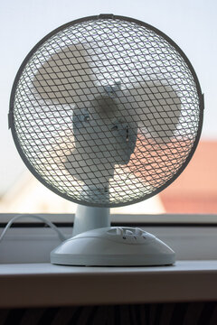 Electric fan blowing air on desk near window in sunny room. Household appliance cooling during extreme heat wave. Summer weather climate control and ventilation equipment concept.