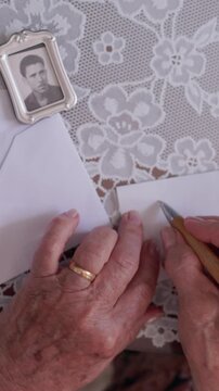 Close up of an elderly woman's wrinkled hands carefully writing a love letter with a wooden pen on a lace tablecloth, next to a small framed photograph of a man from the past