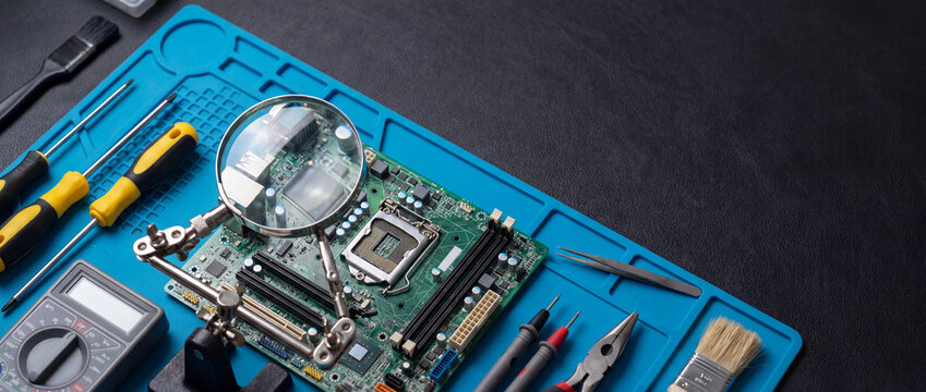 Electronics repair desk flat lay featuring a computer motherboard under a magnifying glass on a blue silicone work mat with screwdrivers, multimeter, and pliers for technical service banners